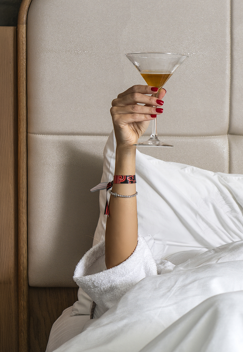 A woman holding a martini glass in a hotel room bed.